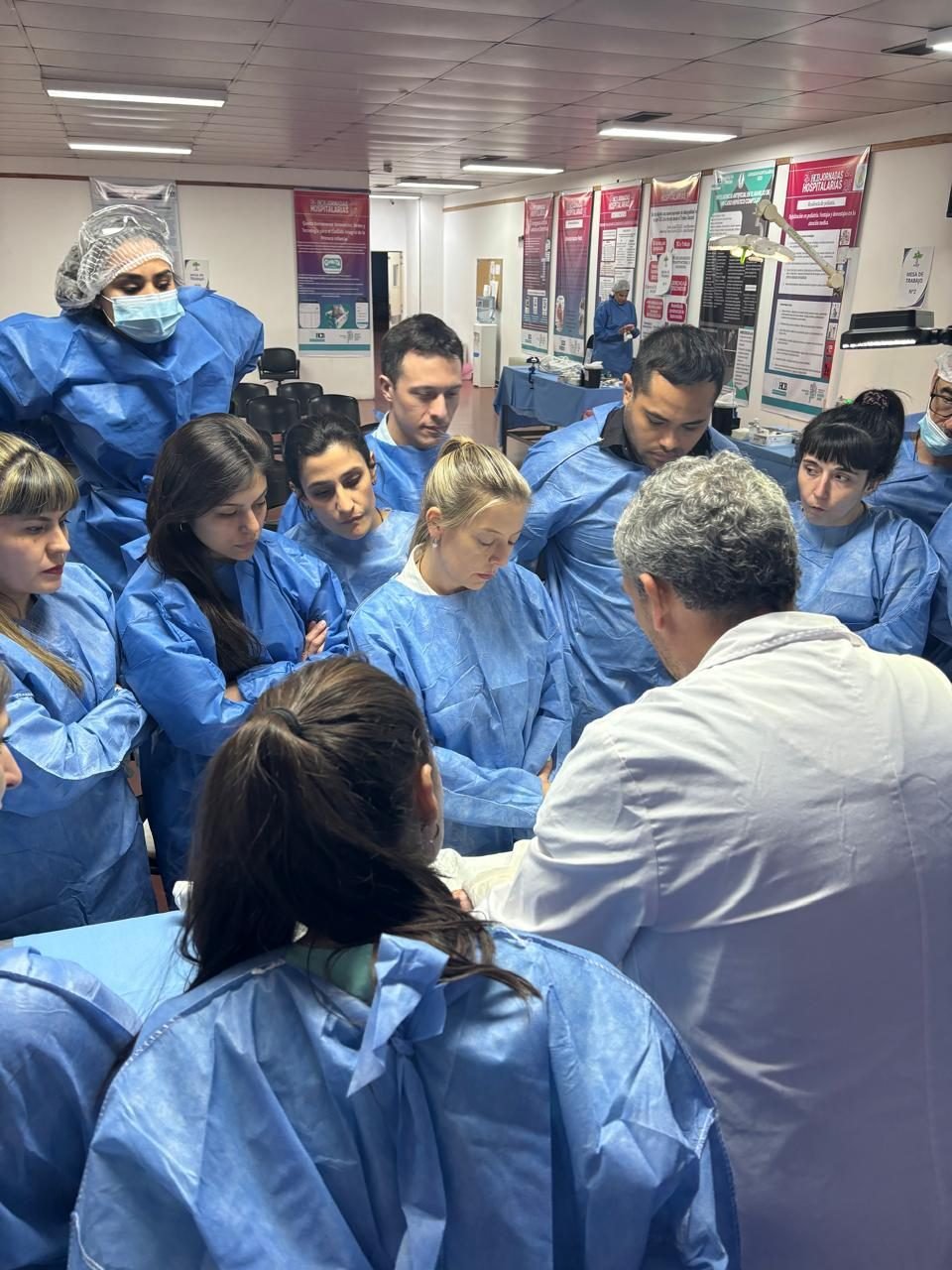Grupo numeroso de participantes con indumentaria quirúrgica rodeando al instructor en la mesa de trabajo